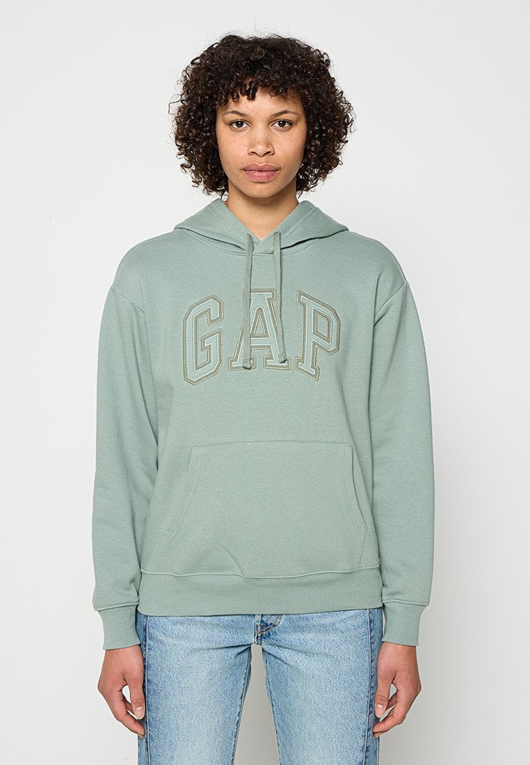Person mit kurzen lockigen Haaren, trägt hellgrünen GAP-Hoodie und blaue Jeans, steht vor weißem Hintergrund.