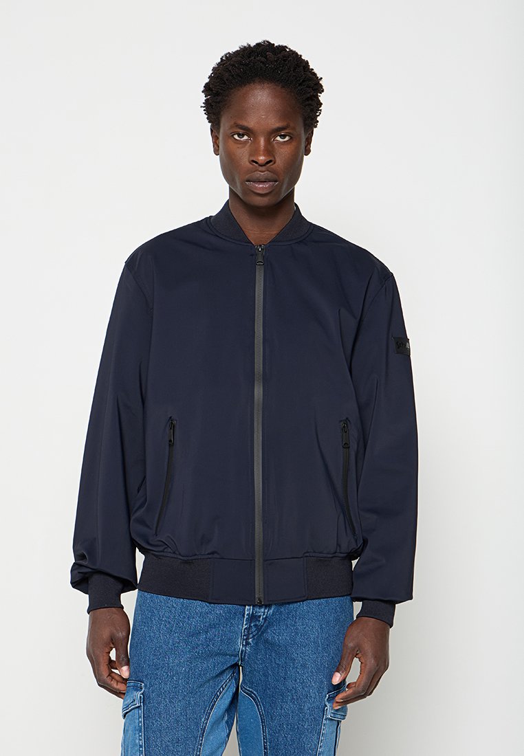 Mann trägt eine marineblaue Bomberjacke mit Reißverschluss und blaue Jeans, steht mit neutralem Gesichtsausdruck vor einem schlichten Hintergrund.