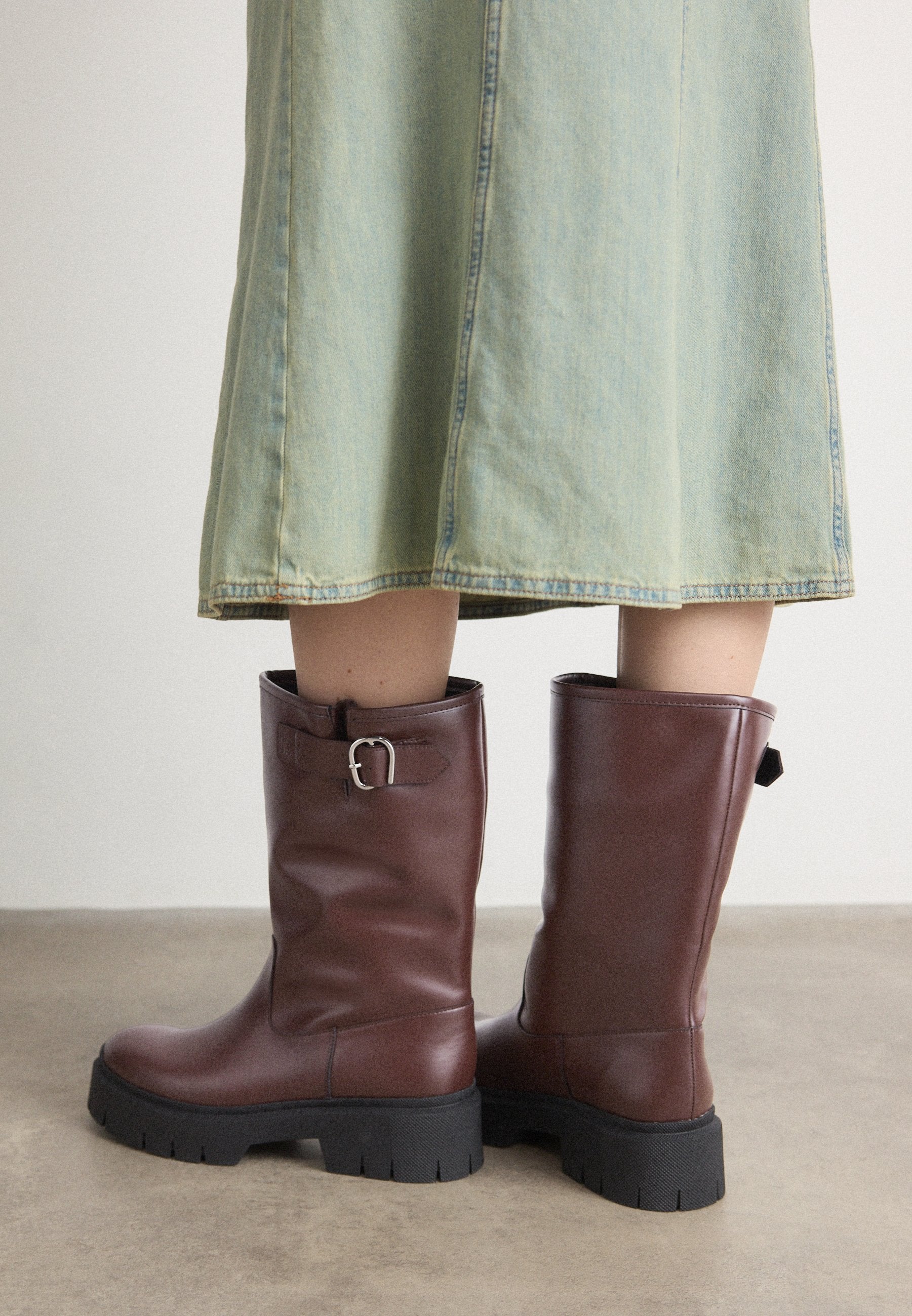 Braune Lederstiefel bis zum Knie mit strukturierter Oberfläche, seitlichem Schnallen-Detail und robusten schwarzen Gummisohlen.