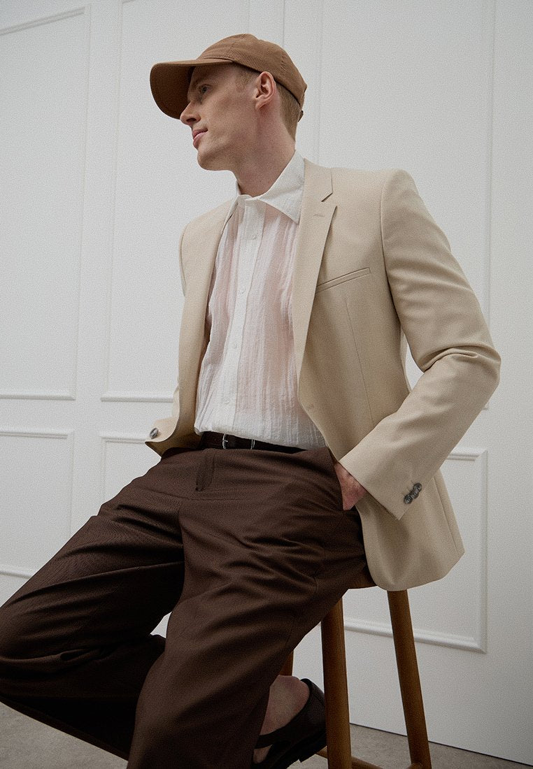 Beiger maßgeschneiderter Blazer über einem halbtransparenten weißen Hemd, braune Hosen und eine beige Kappe, sitzend auf einem Holzschemel vor neutralem Hintergrund.