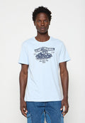 Junger Mann trägt ein hellblaues T-Shirt mit einer schwarzen Vintage-Motorrad-Grafik und Schriftzug, kombiniert mit blauen Cargo-Jeans.