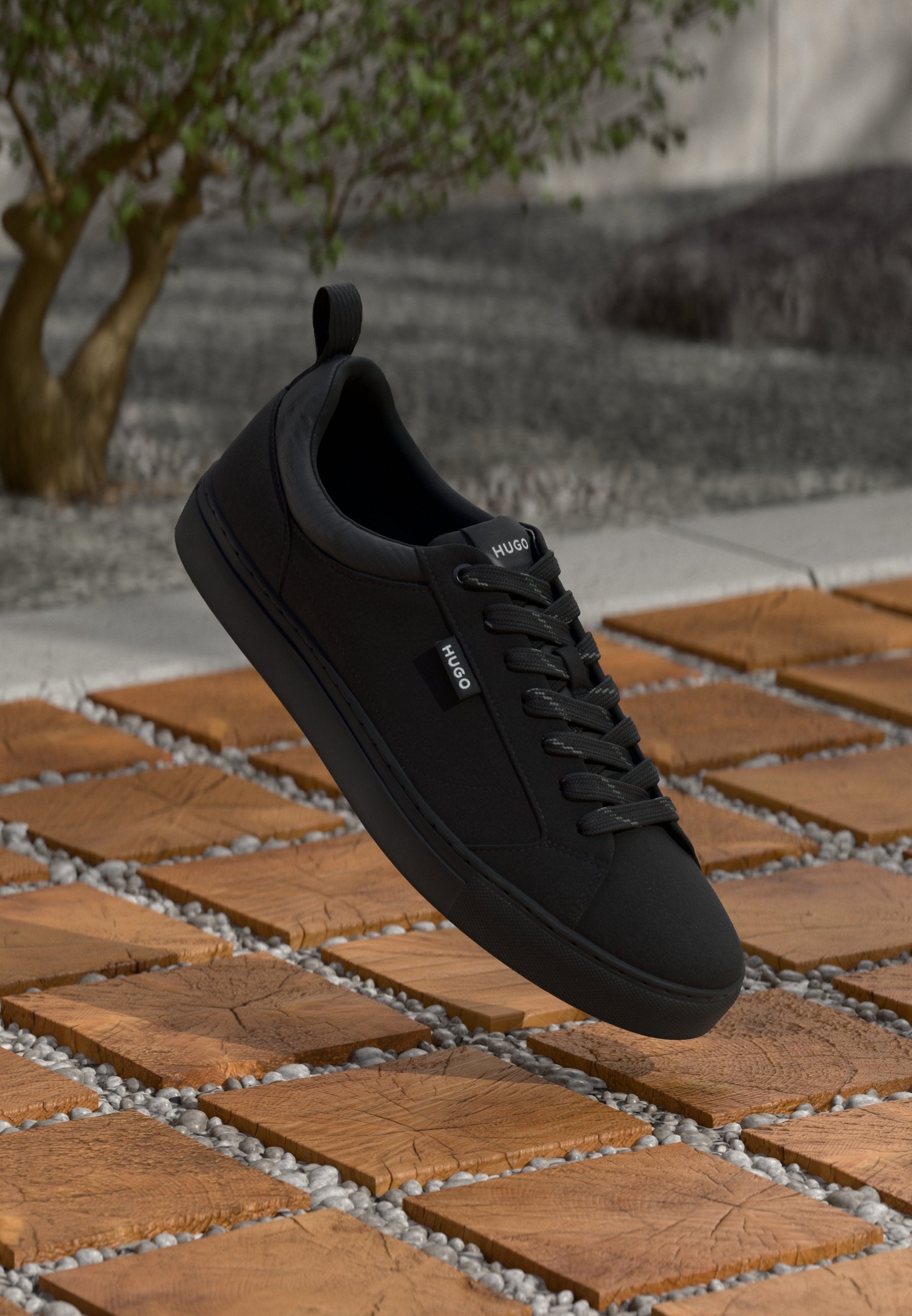 Schwarze Sneaker mit niedrigem Schaft aus strukturiertem Leder, flachen Schnürsenkeln und einem Schlaufenetikett, auf einer gemusterten Fliesenoberfläche mit Kieselsteinen präsentiert.