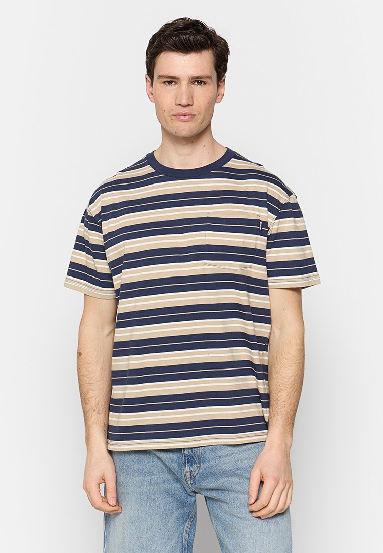 Mann steht da und trägt ein navyblaues, beiges und weiß gestreiftes T-Shirt mit kurzen Ärmeln und einer Brusttasche sowie eine hellblaue Jeans.