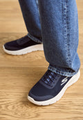 Marineblaue Mesh-Sneaker mit einer weißen Sohle sind mit Schnürsenkeln und einem Logo ausgestattet und werden von hellblauen Jeans und einem Holzboden ergänzt.