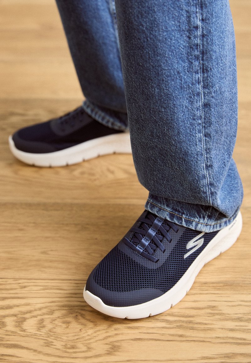 Marineblaue Mesh-Sneaker mit einer weißen Sohle sind mit Schnürsenkeln und einem Logo ausgestattet und werden von hellblauen Jeans und einem Holzboden ergänzt.