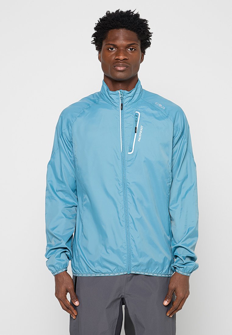Mann steht mit Blick nach vorne, trägt eine hellblaue Zip-Jacke mit einer vertikalen Brusttasche und dunkelgraue Hose.