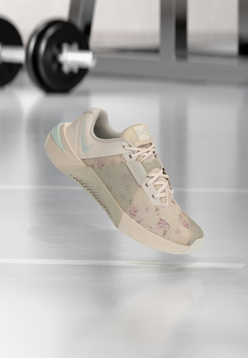 Hellbeiger Sportschuh mit pastellblauen Akzenten und floralem Muster, der über einem reflektierenden Fitnessstudio-Boden schwebt, mit verschwommenen Hanteln im Hintergrund.
