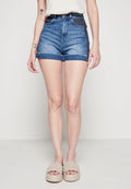 Frau in hochgeschnittenen blauen Denim-Shorts mit gemusterten Seitenpaneelen, beigen geflochtenen Plateausandalen und weißem ärmellosem Oberteil, stehend.