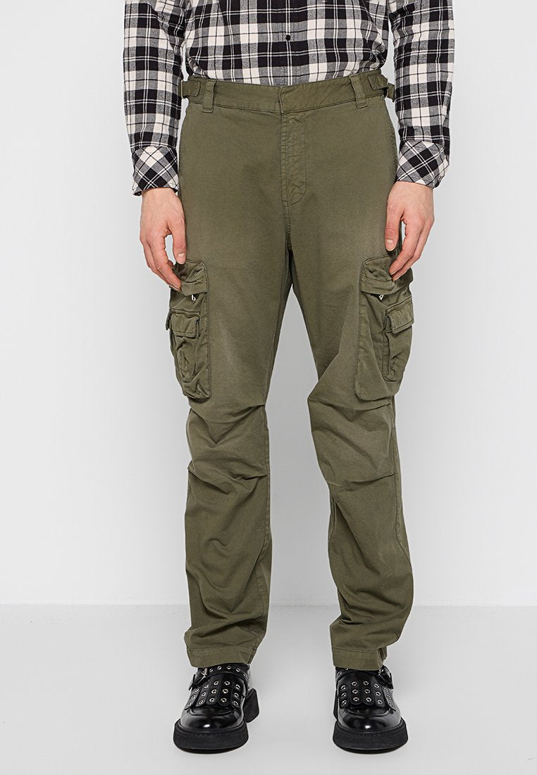 Cargo-Hosen aus einem olivgrünen Baumwollmix, ausgestattet mit mehreren Reißverschluss- und Klappentaschen, lockerem Schnitt und verstärkten Knien. Schwarze Schuhe mit Nieten.