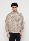 Mann mit lockigem Haar, der einen beige-schwarzen Leopardenmuster-Hoodie und schwarze Hosen trägt, steht vor einem schlichten hellen Hintergrund.