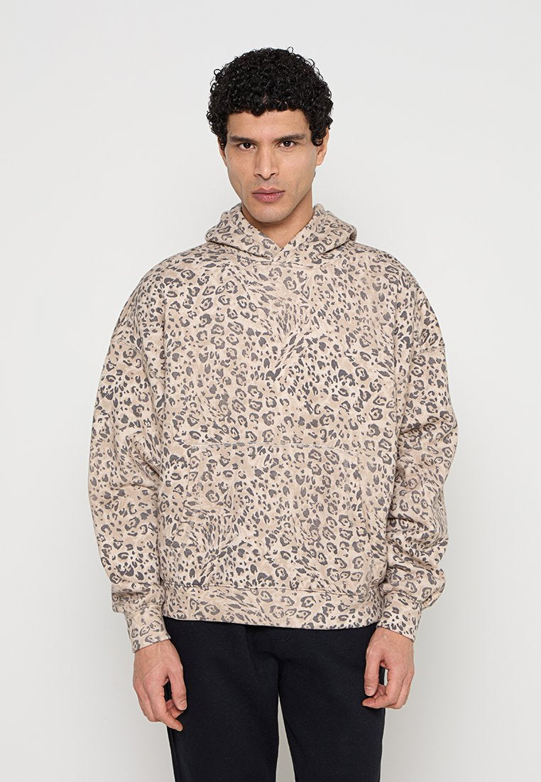 Mann mit lockigem Haar, der einen beige-schwarzen Leopardenmuster-Hoodie und schwarze Hosen trägt, steht vor einem schlichten hellen Hintergrund.