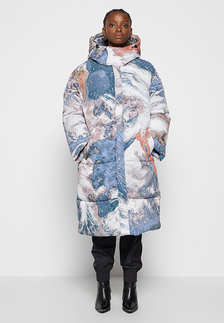 Gepolsterter Mantel mit einem blauen, weißen und rosa abstrakten Muster, mit hohem Kragen, Kapuze und Vordertaschen. Getragen über schwarzen Hosen und Stiefeln.