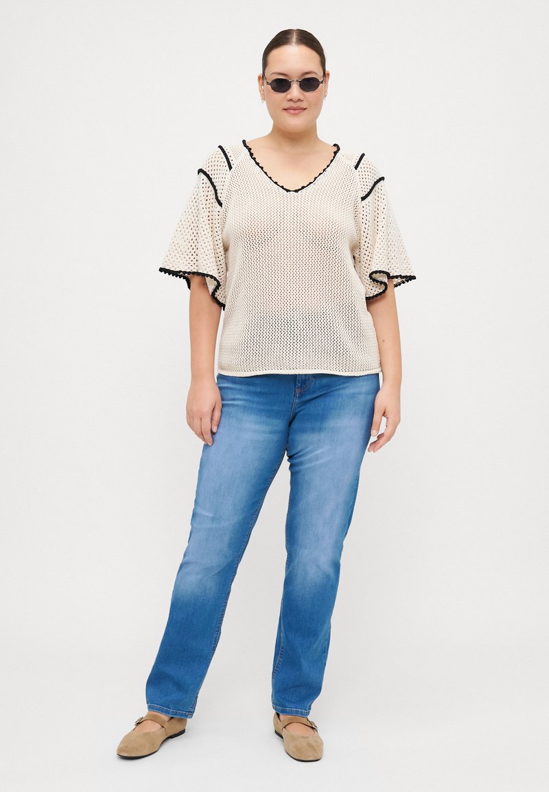 Beige gestricktes Oberteil mit weiten Ärmeln, schwarz abgesetzt, kombiniert mit hellblauen geraden Jeans und beigen Schuhen. Glatte Textur, offene Maschen.