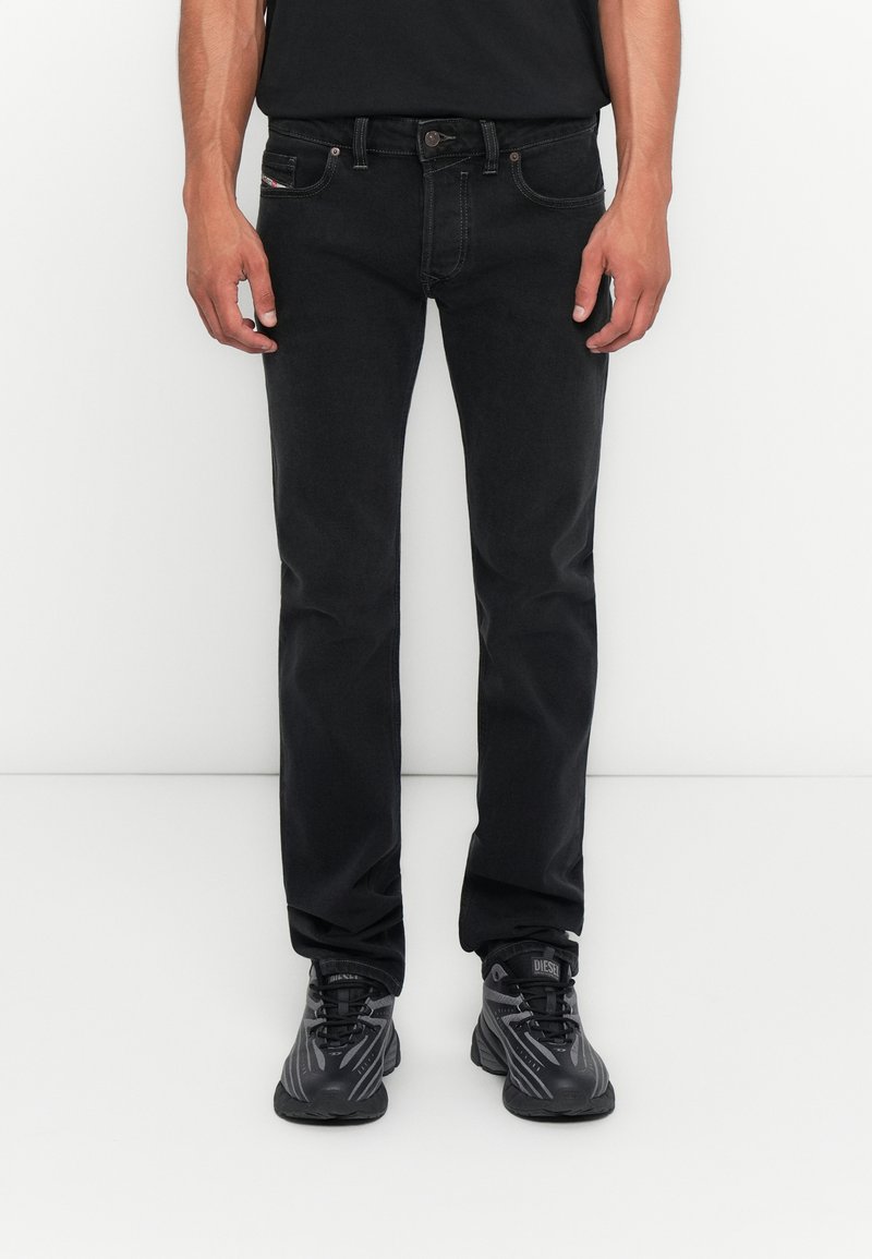 Mann in engen schwarzen Jeans und schwarzen Sport Schuhen, der vor einem weißen Hintergrund steht, die Hände entspannt an den Seiten.