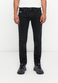 Mann, der enge schwarze Jeans, ein weißes Hemd und schwarze Sneakers trägt, steht vor einem schlichten weißen Hintergrund.