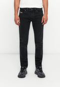 Mann, der enge schwarze Jeans, ein weißes Hemd und schwarze Sneakers trägt, steht vor einem schlichten weißen Hintergrund.