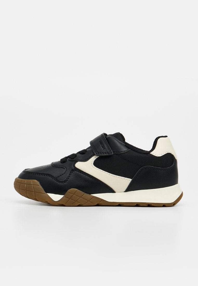 Schwarz-weißer Sneaker mit brauner Gummisohle, Schnürung vorne und einem schwarzen Klettverschluss oben, von der Seite gezeigt.