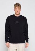 Mann trägt ein schwarzes Sweatshirt mit langen Ärmeln und kleinem weißem "AllSaints London"-Logo vorne, beige Hose, steht vor einfachem Hintergrund.