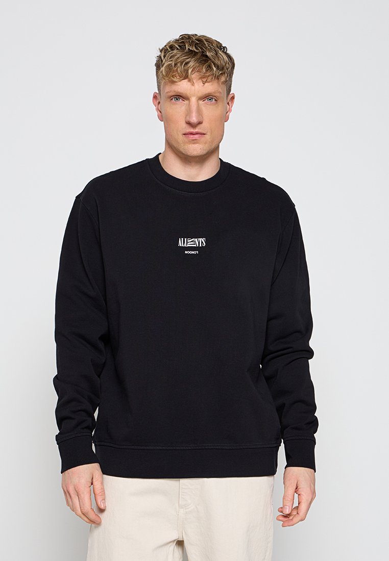 Mann trägt ein schwarzes Sweatshirt mit langen Ärmeln und kleinem weißem "AllSaints London"-Logo vorne, beige Hose, steht vor einfachem Hintergrund.