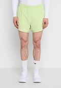 Mann, der hellgrüne Sportshorts, ein weißes Langarmshirt, weiße Sportsocken und weiße Turnschuhe trägt und vor einem einfarbigen Hintergrund steht.