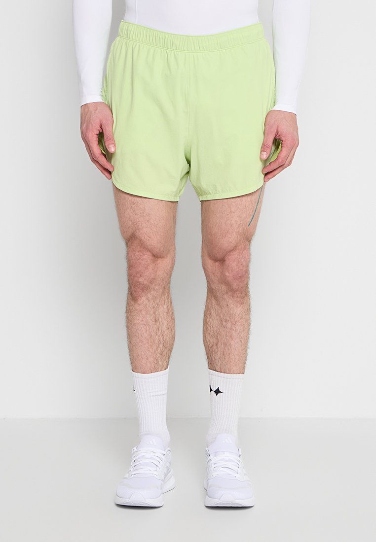 Mann, der hellgrüne Sportshorts, ein weißes Langarmshirt, weiße Sportsocken und weiße Turnschuhe trägt und vor einem einfarbigen Hintergrund steht.