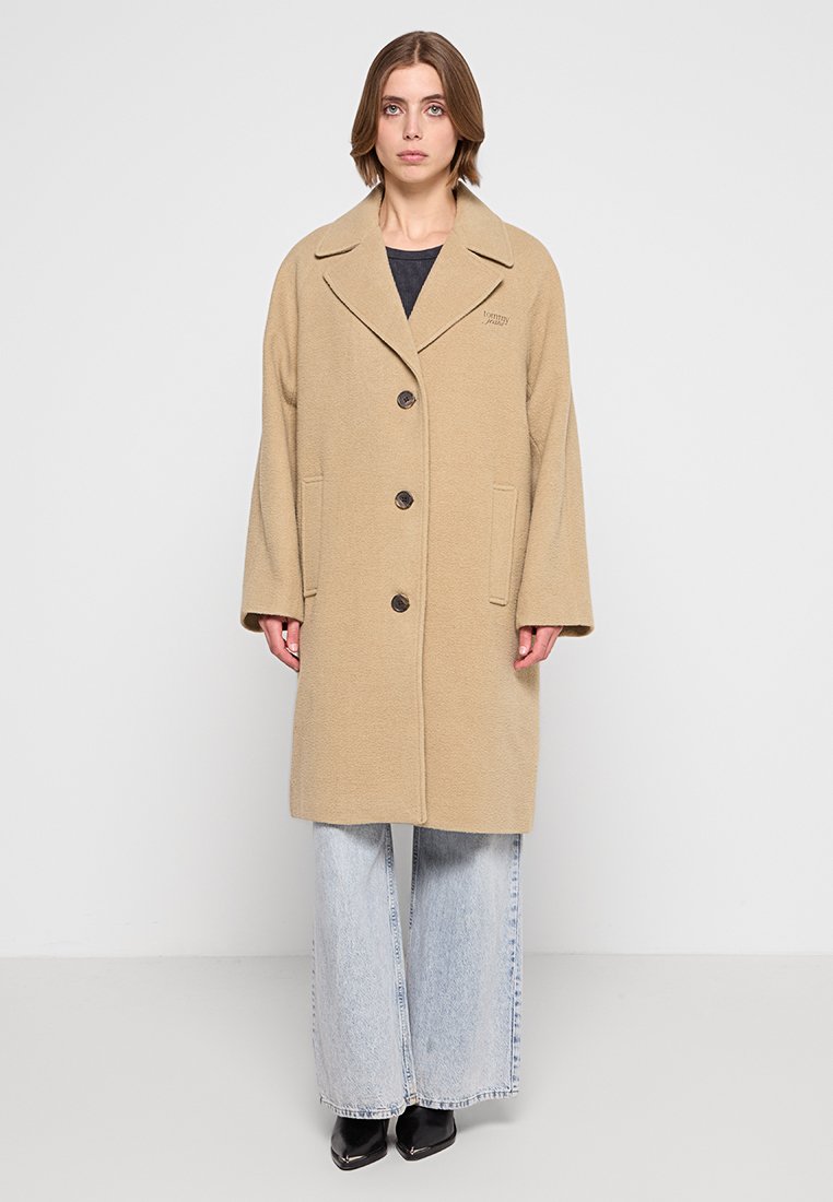 Beige Wollmantel mit Reverskragen, vier Knöpfen und Seitentaschen, kombiniert mit lockeren hellblauen Jeans und schwarzen Stiefeln.