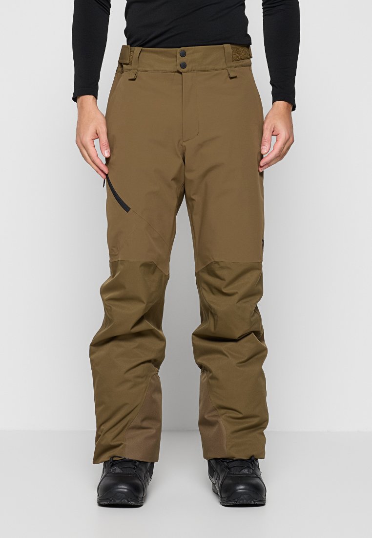 Mann trägt braune Outdoor-Hose mit verstärkten Knien und schwarze Wanderschuhe, steht mit entspannt an den Seiten hängenden Händen.