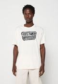 Eine dunkelhäutige Person trägt ein off-white O’Neill T-Shirt mit grauem Camouflage-Logo und hellbeigen Hosen und steht vor einem schlichten Hintergrund.