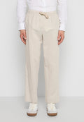 Mann trägt beige Leinenhose mit Tunnelzug, weißes Hemd mit Knöpfen und weiße Turnschuhe, steht vor einem schlichten Hintergrund.