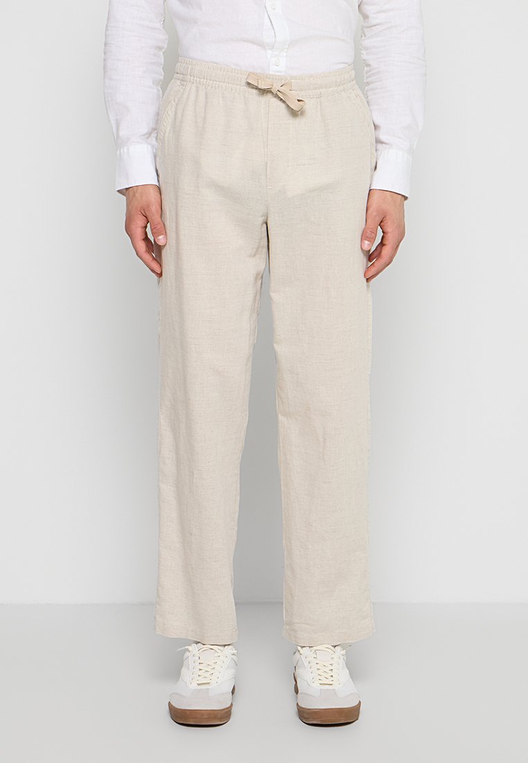 Mann trägt beige Leinenhose mit Tunnelzug, weißes Hemd mit Knöpfen und weiße Turnschuhe, steht vor einem schlichten Hintergrund.