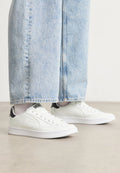 Weiße Sneaker mit einer glatten Ledertextur, schwarzen Fersenakzenten und perforierten Details. Kombiniert mit hellblauen, weiten Jeanshosen.