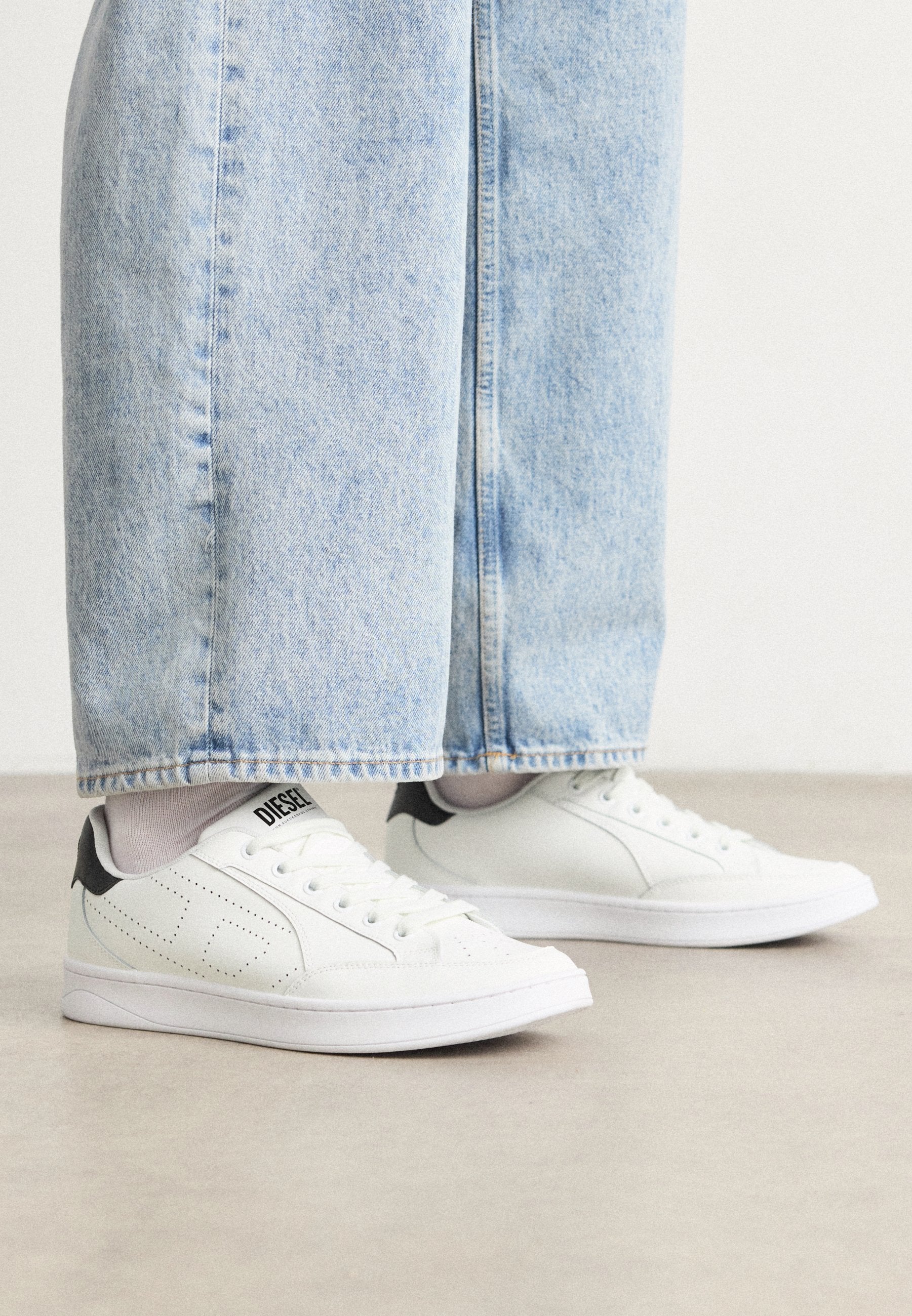Weiße Sneaker mit einer glatten Ledertextur, schwarzen Fersenakzenten und perforierten Details. Kombiniert mit hellblauen, weiten Jeanshosen.