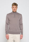 Mann trägt hellbraunen Zip-up-Pullover mit hohem Kragen und beiger Hose, steht vor einem einfarbigen hellen Hintergrund.