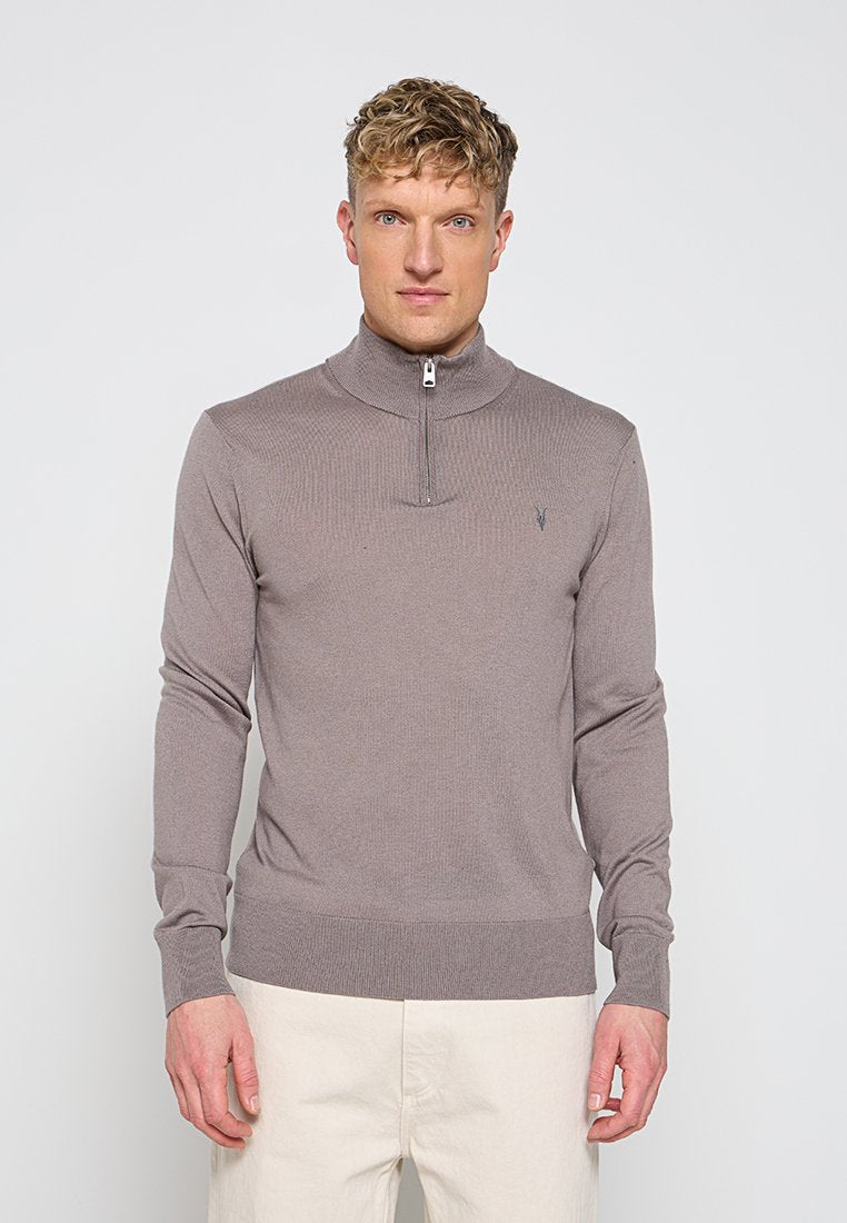 Mann trägt hellbraunen Zip-up-Pullover mit hohem Kragen und beiger Hose, steht vor einem einfarbigen hellen Hintergrund.