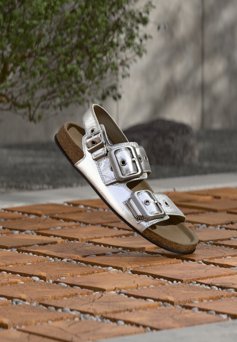 Silberne, offene Sandale mit Metallic-Finish, zwei verstellbaren Riemen mit Schnallen, auf einem Boden aus Holz und Kieselsteinen im Freien liegend.