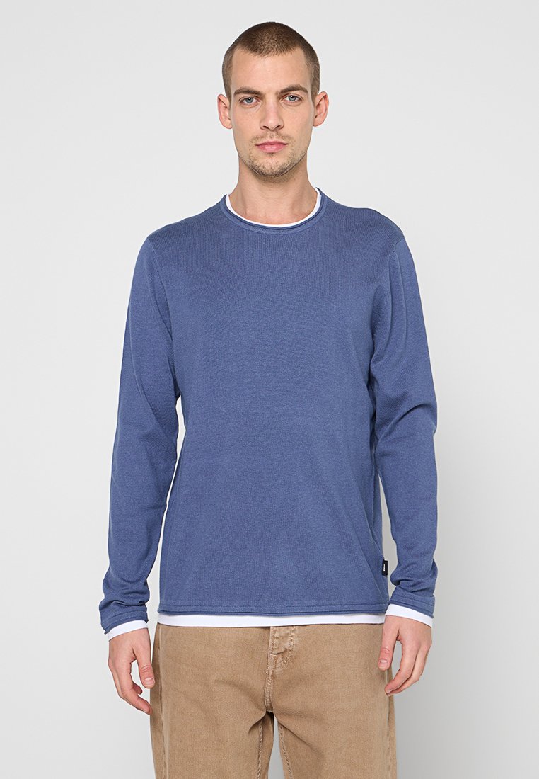 Marineblauer Pullover mit langen Ärmeln, strukturierter Oberfläche und weißem Besatz am Hals, kombiniert mit hellbraunen Hosen.