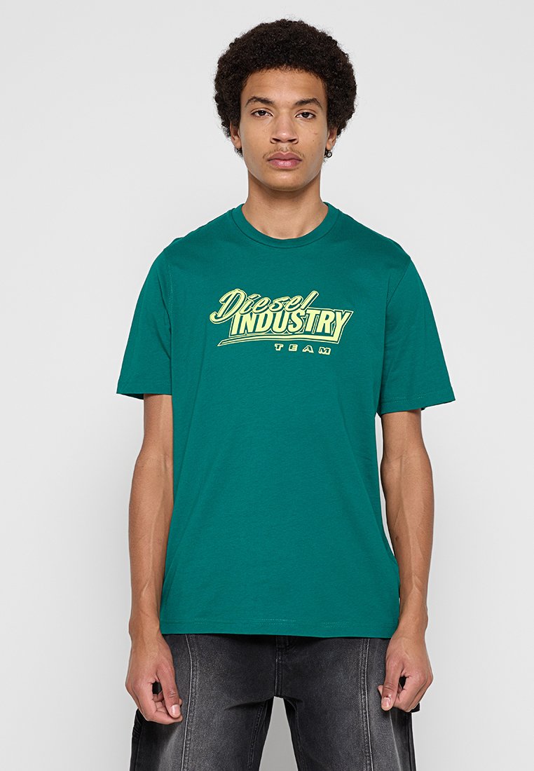 Junger Mann steht mit Blick nach vorne, trägt ein grünes Diesel Industry Team T-Shirt und dunkle Jeans vor einem schlichten Hintergrund.