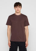 Junger Mann, der nach vorne blickt, trägt ein einfarbiges braunes Kurzarm-T-Shirt und schwarze Hose vor grauem Hintergrund.