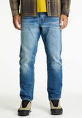 Mann in ausgewaschenen blauen Jeans, beigen und schwarzen Sneakern, gelbem Hemd und braunem kariertem Jackett, das vor einem weißen Hintergrund steht.