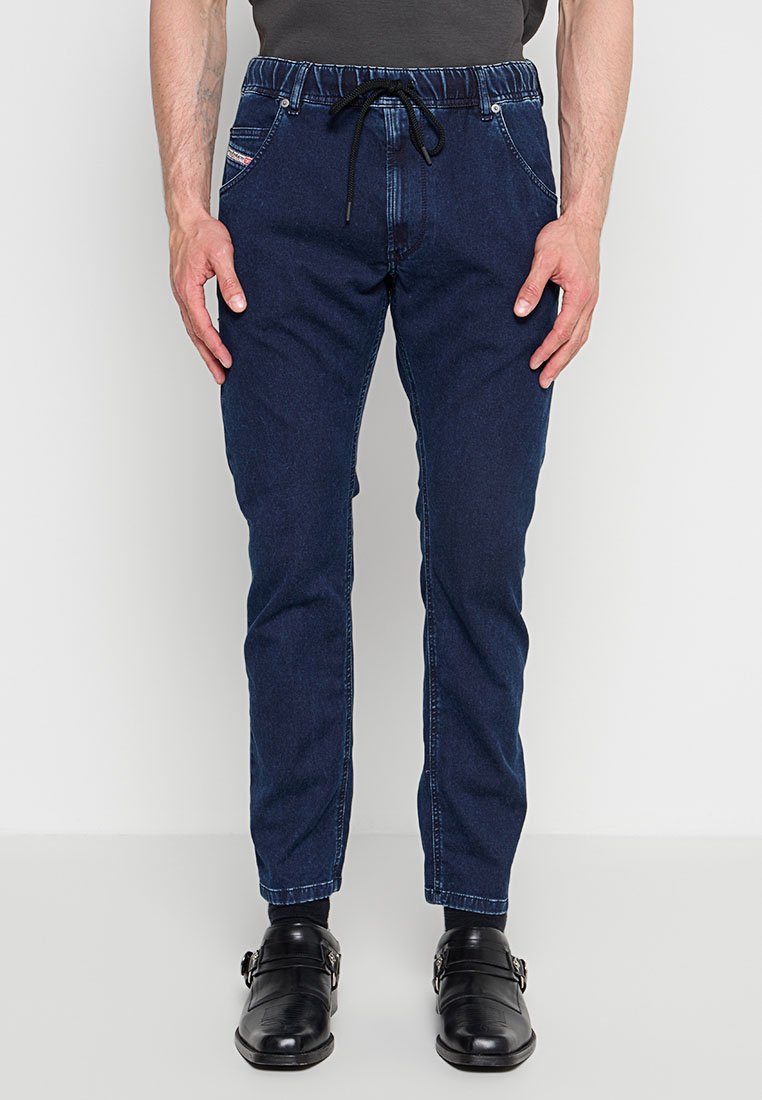 Mann trägt dunkelblaue Denim-Joggerhosen mit elastischem Bund und schwarzen Anzugschuhen, steht vor weißem Hintergrund.