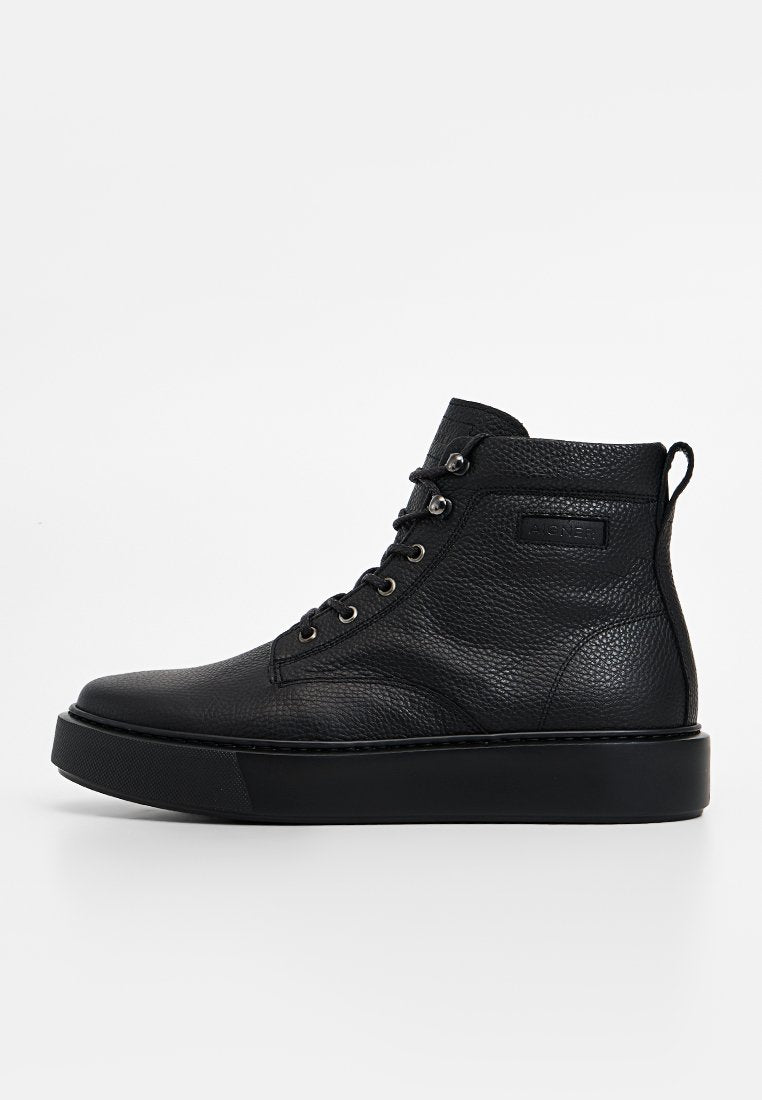 Schwarze High-Top-Schuhe aus Leder mit strukturierter Oberfläche, flacher schwarzer Sohle und Metallösen. Auf der Außenseite befindet sich ein Etikett.