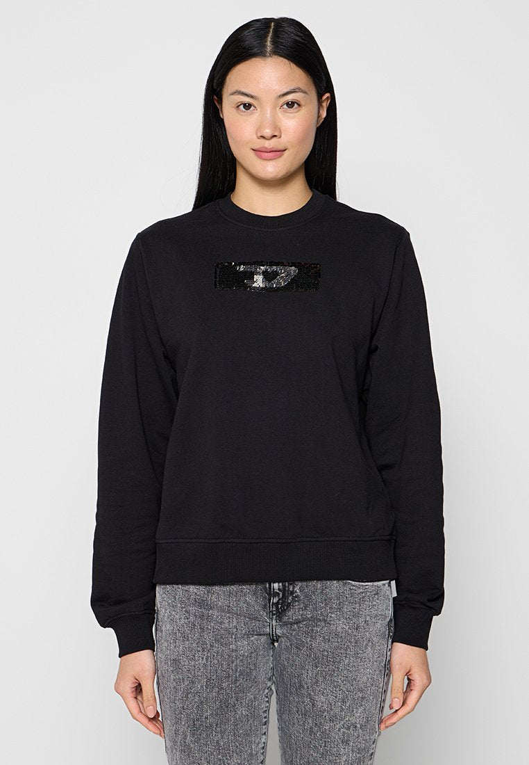 Schwarzes Sweatshirt mit Rundhalsausschnitt und langen Ärmeln, versehen mit einem rechteckigen Pailletten-Patch auf der Vorderseite. Getragen mit grauen Jeans.