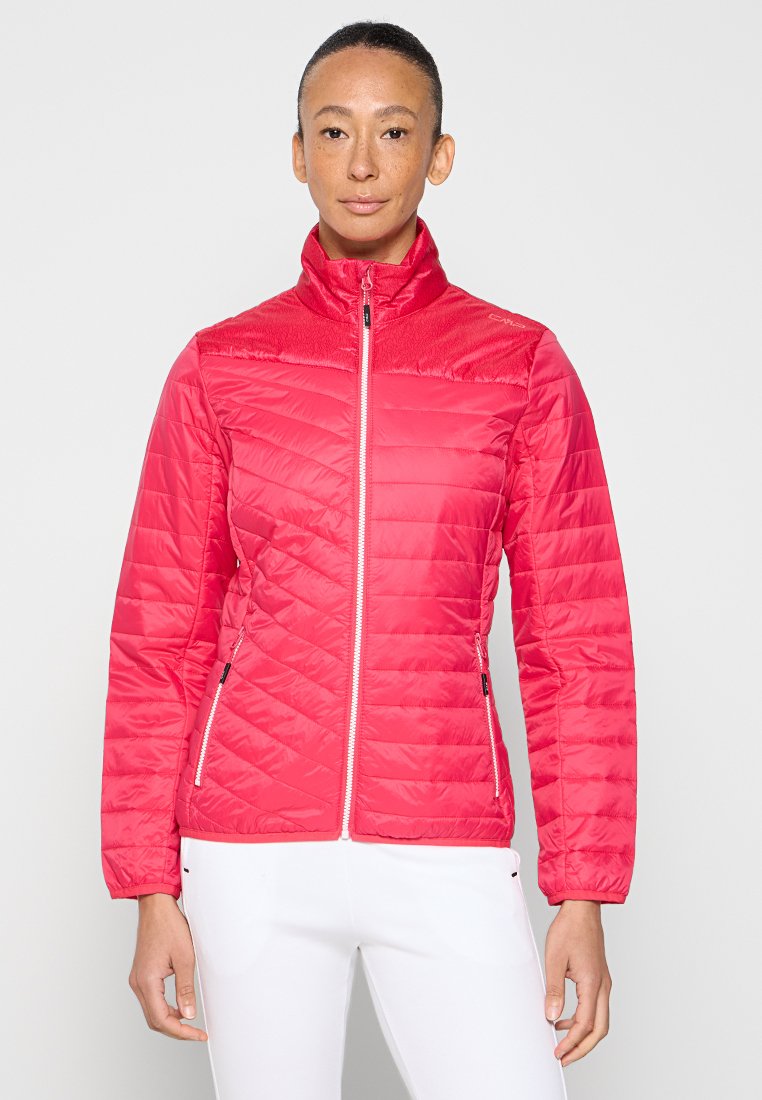 Frau trägt eine leuchtend pinke gesteppte Jacke mit weißen Reißverschlusstaschen und steht vor einem schlichten hellen Hintergrund.