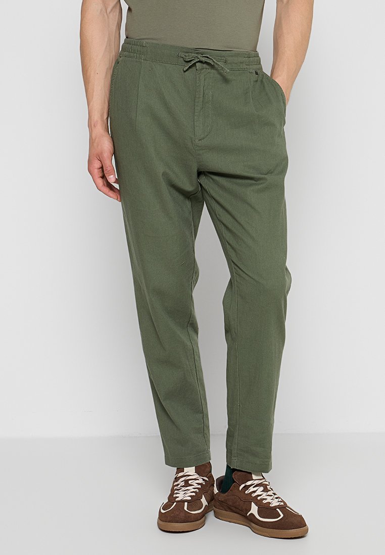 Mann trägt olivgrüne Hose mit Kordelzug und braune Sneakers, steht vor schlichtem grauem Hintergrund.