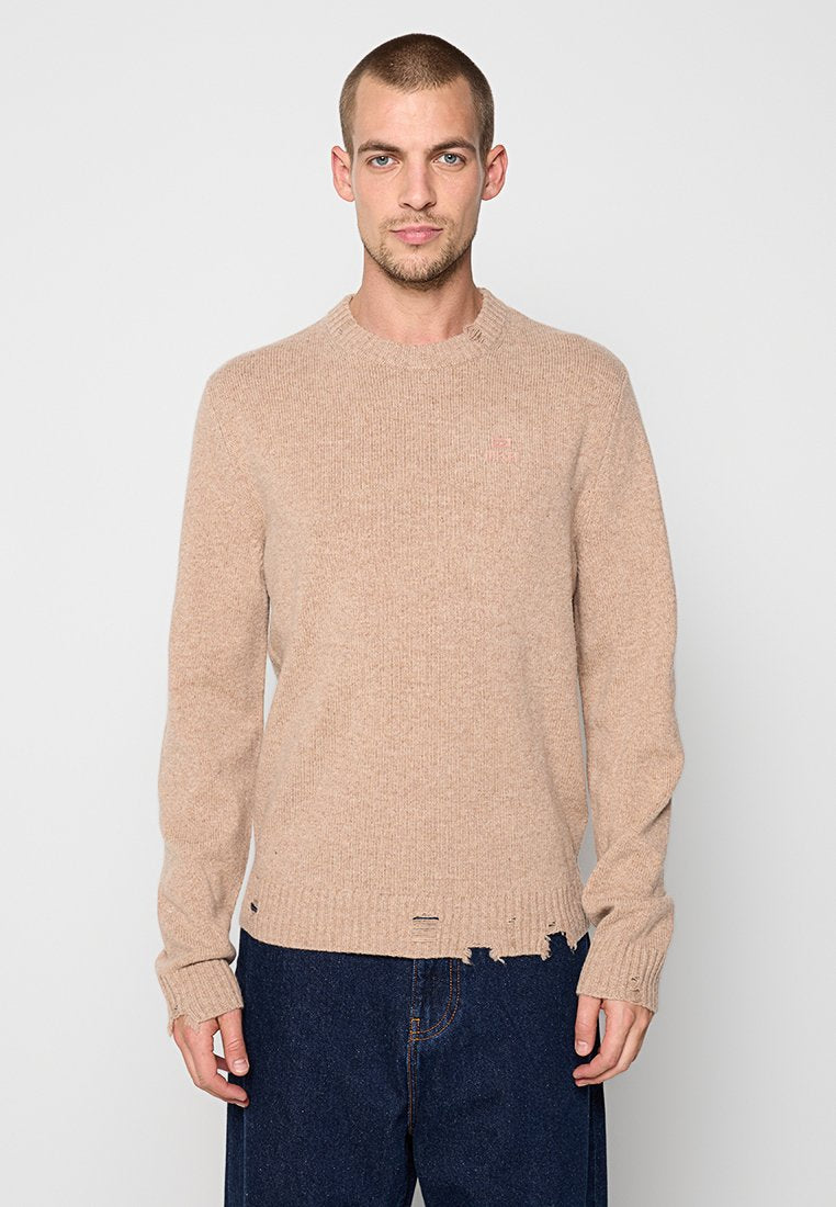 Mann mit kurzen Haaren, der einen beigen Strickpullover mit ausgefransten Säumen und Bündchen trägt, kombiniert mit dunkelblauen Jeans, steht frontal.