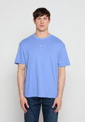 Mann trägt ein hellblaues Tommy Jeans T-Shirt und dunkelblaue Jeans, steht vor einem einfarbigen hellgrauen Hintergrund.