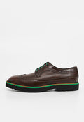 Braune Brogue-Lederschuhe mit dekorativen Perforationen, grünen Schnürsenkeln und einer kontrastierenden grünen und braunen Sohle. Flaches Profil und strukturiertes Design.