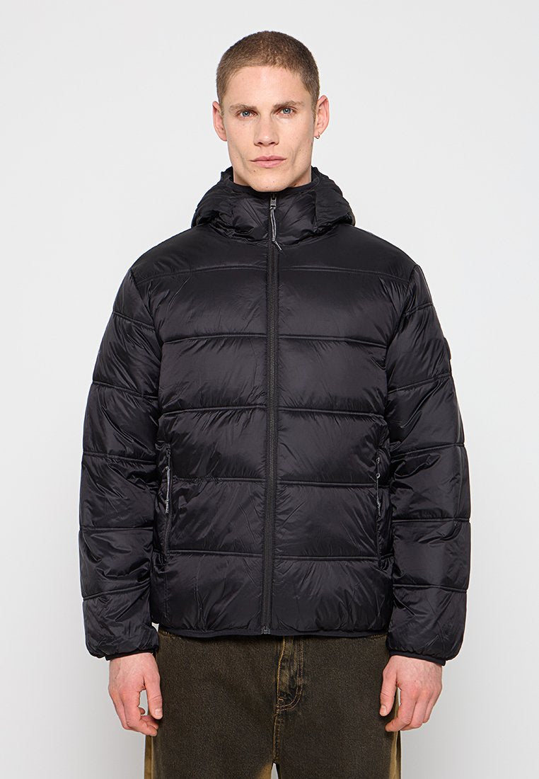 Mann trägt eine schwarze Steppjacke mit Kapuze, die hochgezippt ist, und steht vor einem einfarbigen hellen Hintergrund.