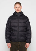 Mann trägt eine schwarze Steppjacke mit Kapuze, die hochgezippt ist, und steht vor einem einfarbigen hellen Hintergrund.