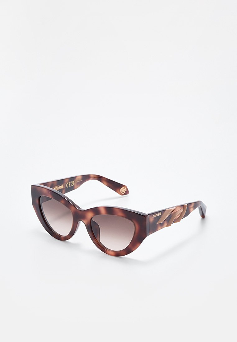 Sonnenbrille im Cat-Eye-Stil mit Schildpattmuster, verlaufend braunen Gläsern und kantigen goldfarbenen Details an den Bügeln, auf einer weißen Oberfläche präsentiert.